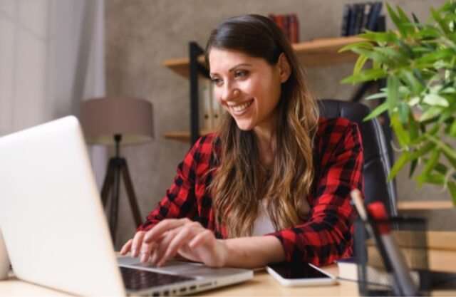 Image of a woman working at a laptop, she is wearing a red and black checked shirt and is seated next to a pot plant
